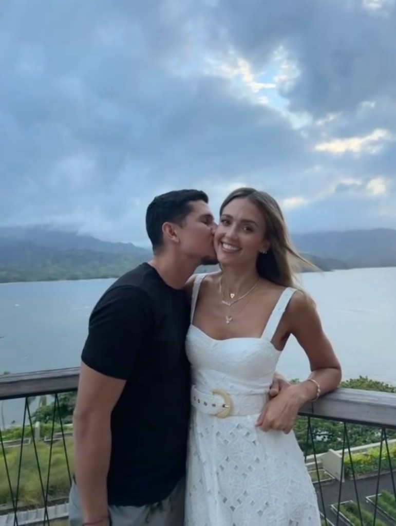 Jessica Alba being kissed on the cheek by a man, standing on a balcony overlooking water and mountains.