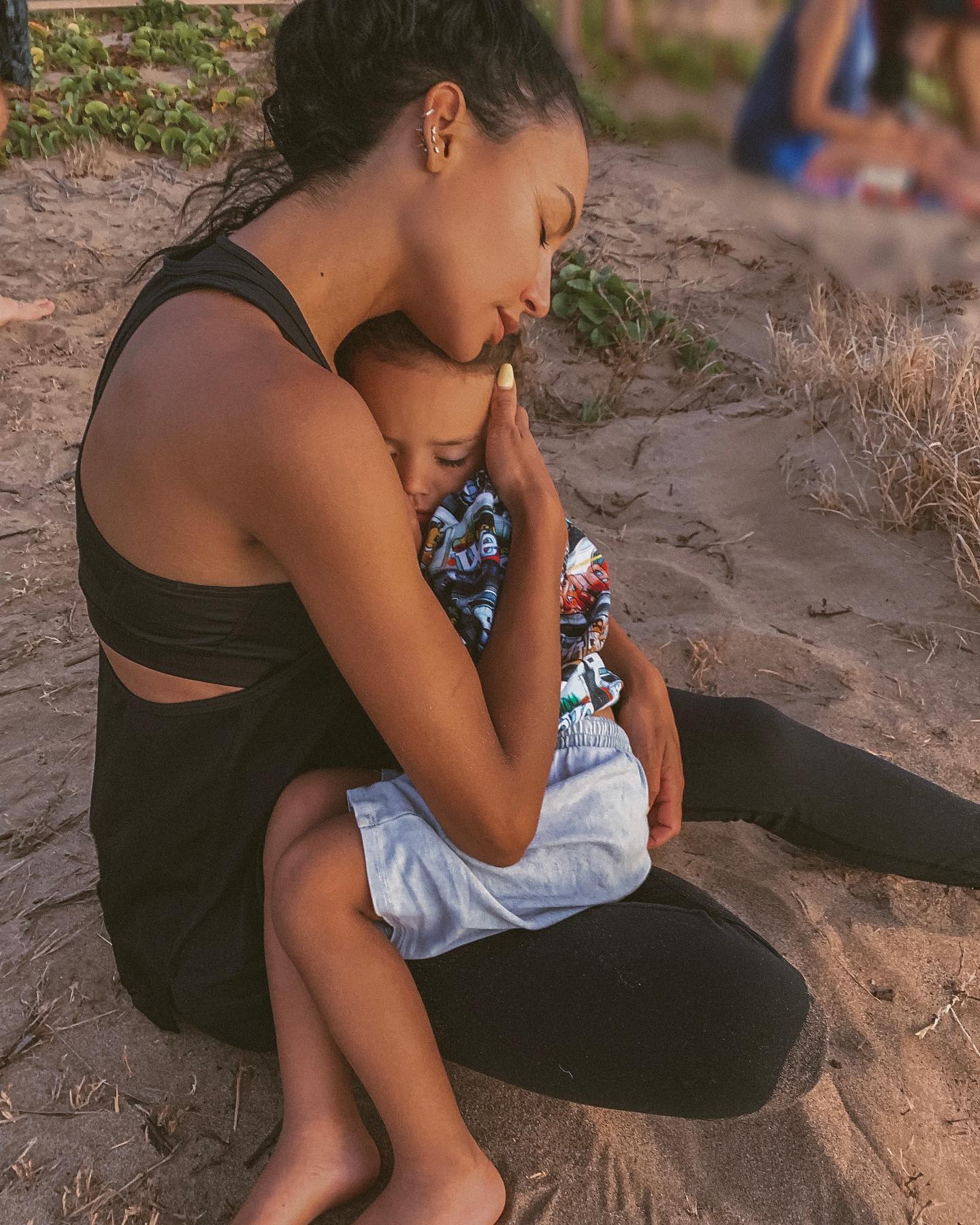 Naya Rivera n a black top hugging her child, who is wearing a colorful shirt and light blue shorts, while sitting on a sandy ground.