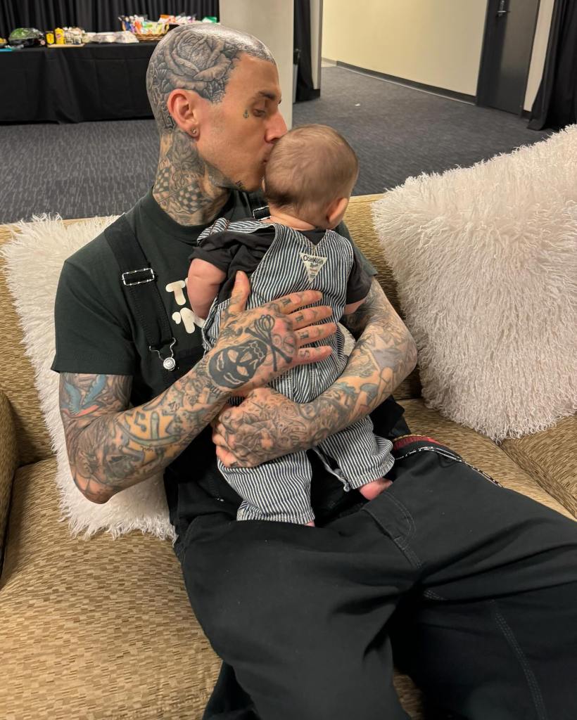 Travis Barker, heavily tattooed, sits on a couch holding and kissing a baby on its forehead.