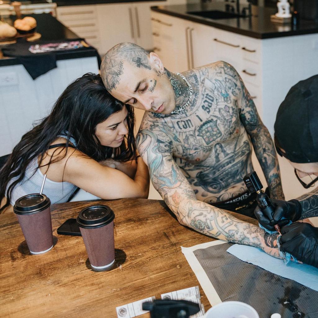 A heavily tattooed man getting a tattoo on his forearm while a woman rests her head on his shoulder.