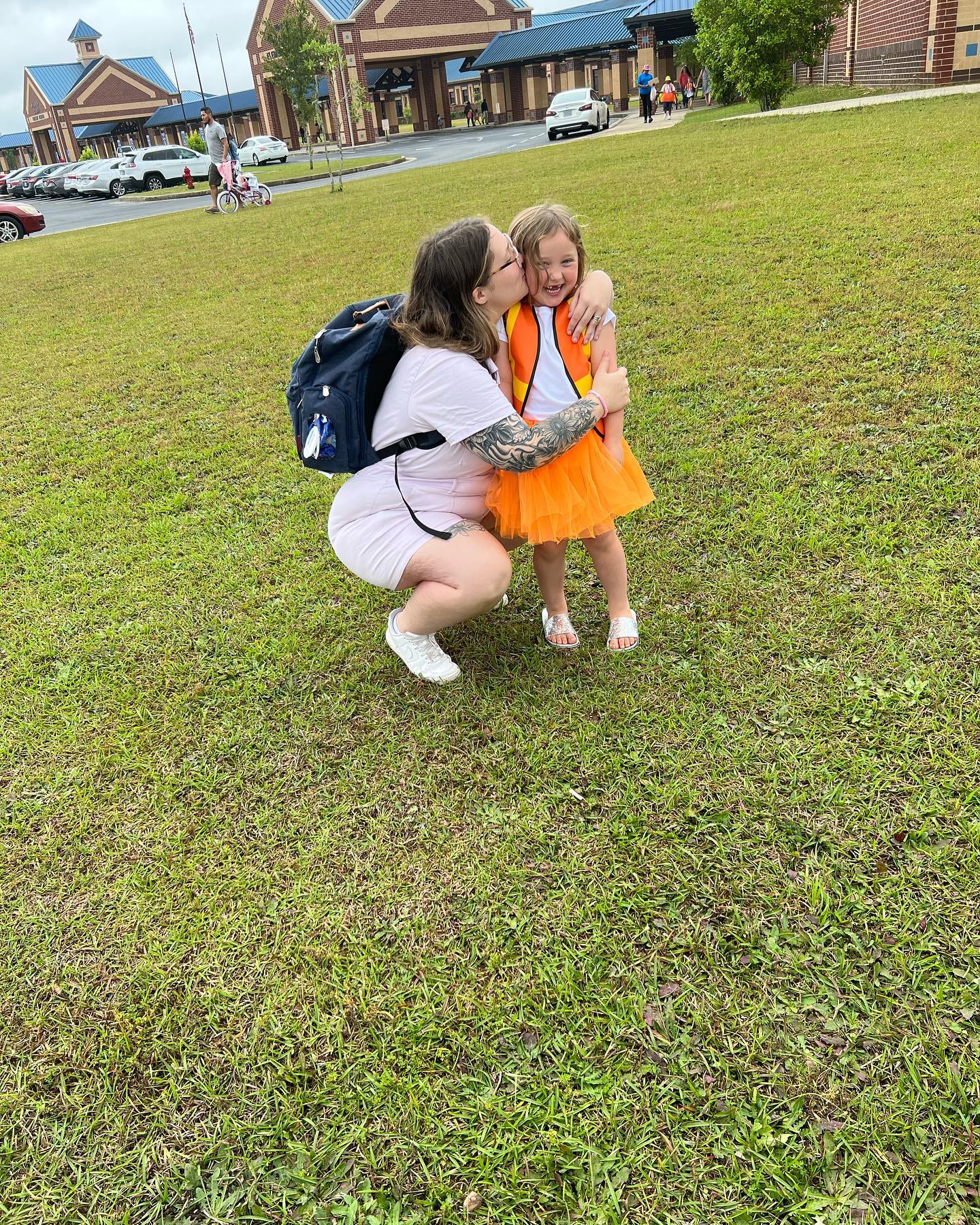 A woman in a white shirt and shorts with a backpack, squatting to kiss a young girl in an orange dress on the cheek.