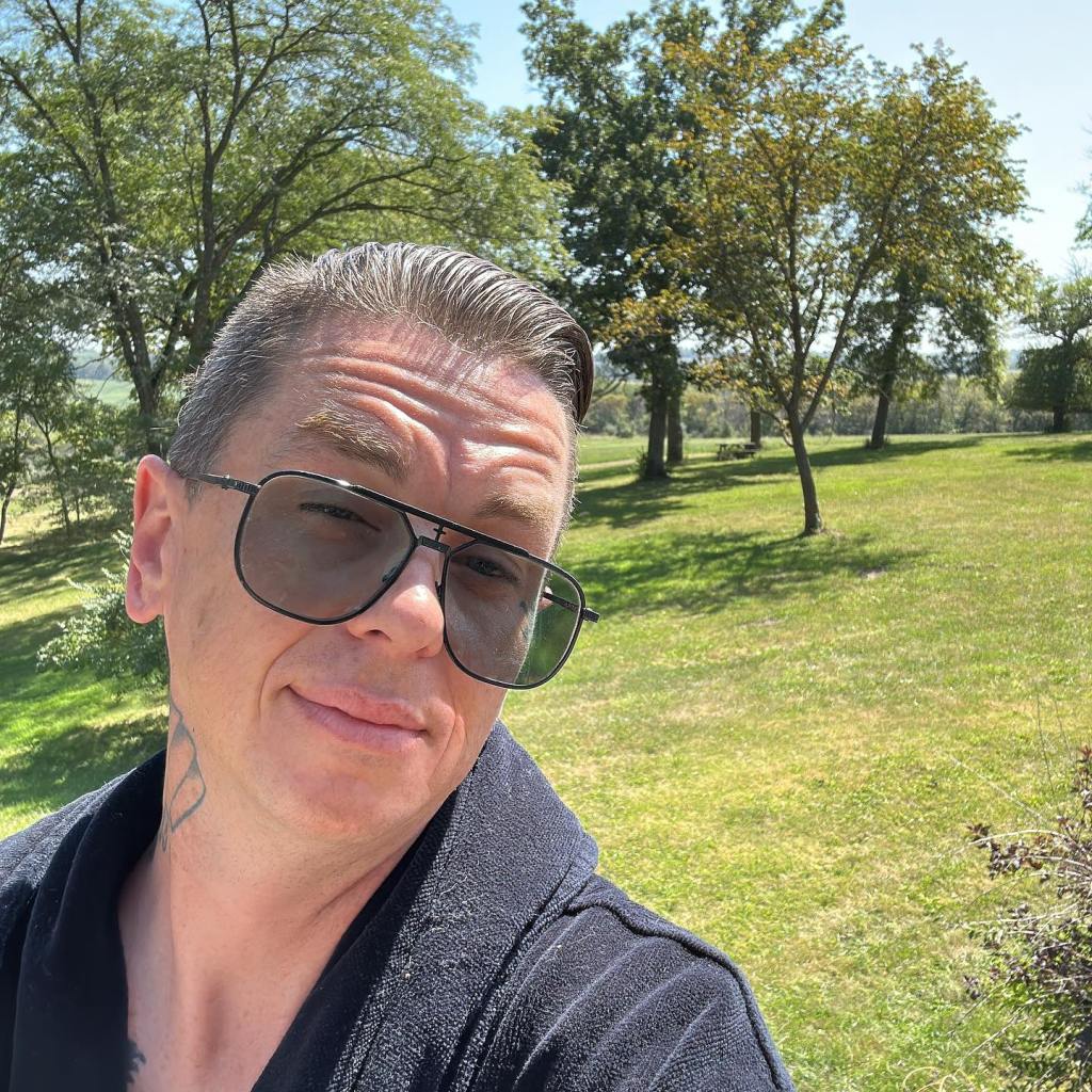 Sid Wilson wearing sunglasses and a black robe outdoors in a field.