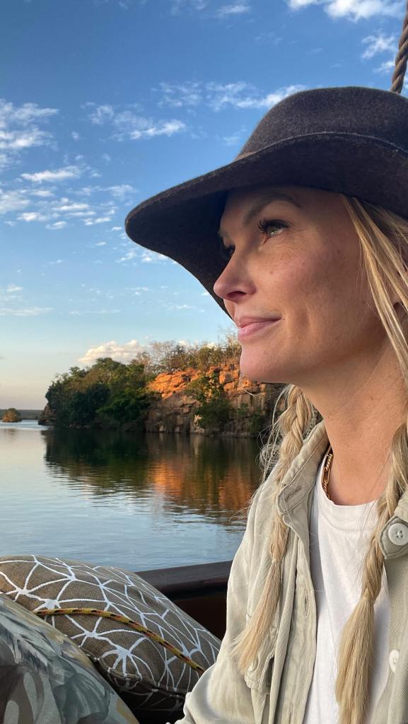 A woman with a brown hat and braided blonde hair looks out at a river with rocky, tree-lined shores under a blue sky.