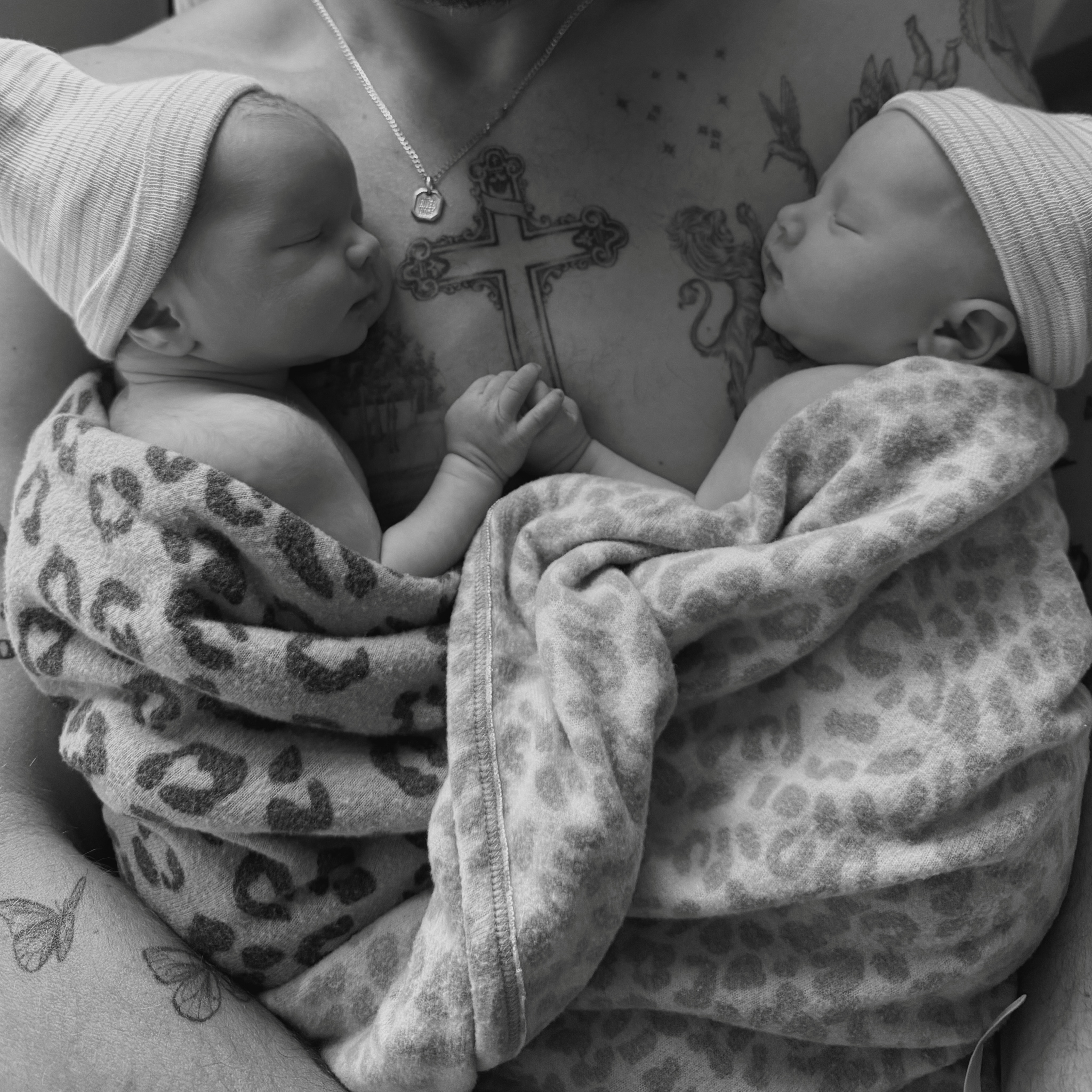 Two swaddled newborn babies in hats resting on a person's tattooed chest.
