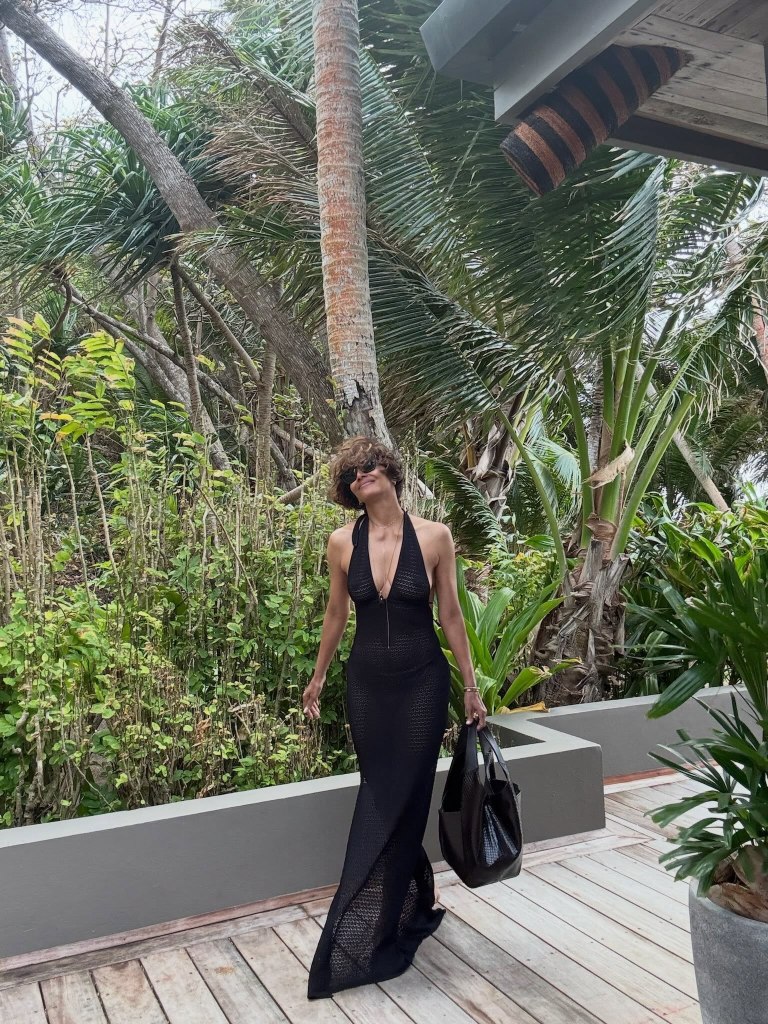 Halle Berry in a black dress, holding a black bag, on a wooden deck surrounded by tropical plants.