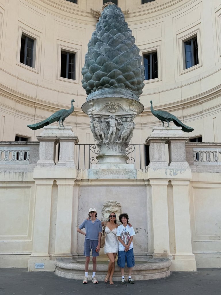 Jennifer Lopez and her twins pose in front of a large pinecone statue and two peacock statues.