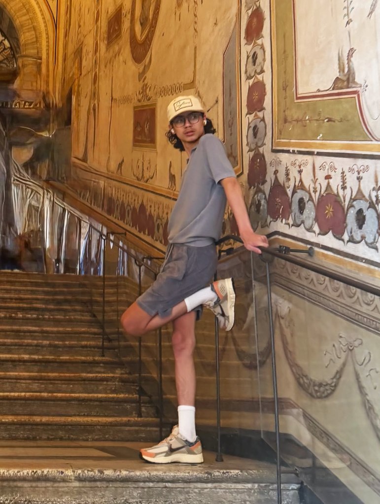 A young man in a t-shirt, shorts, and cap poses on a staircase with frescoed walls in Italy.