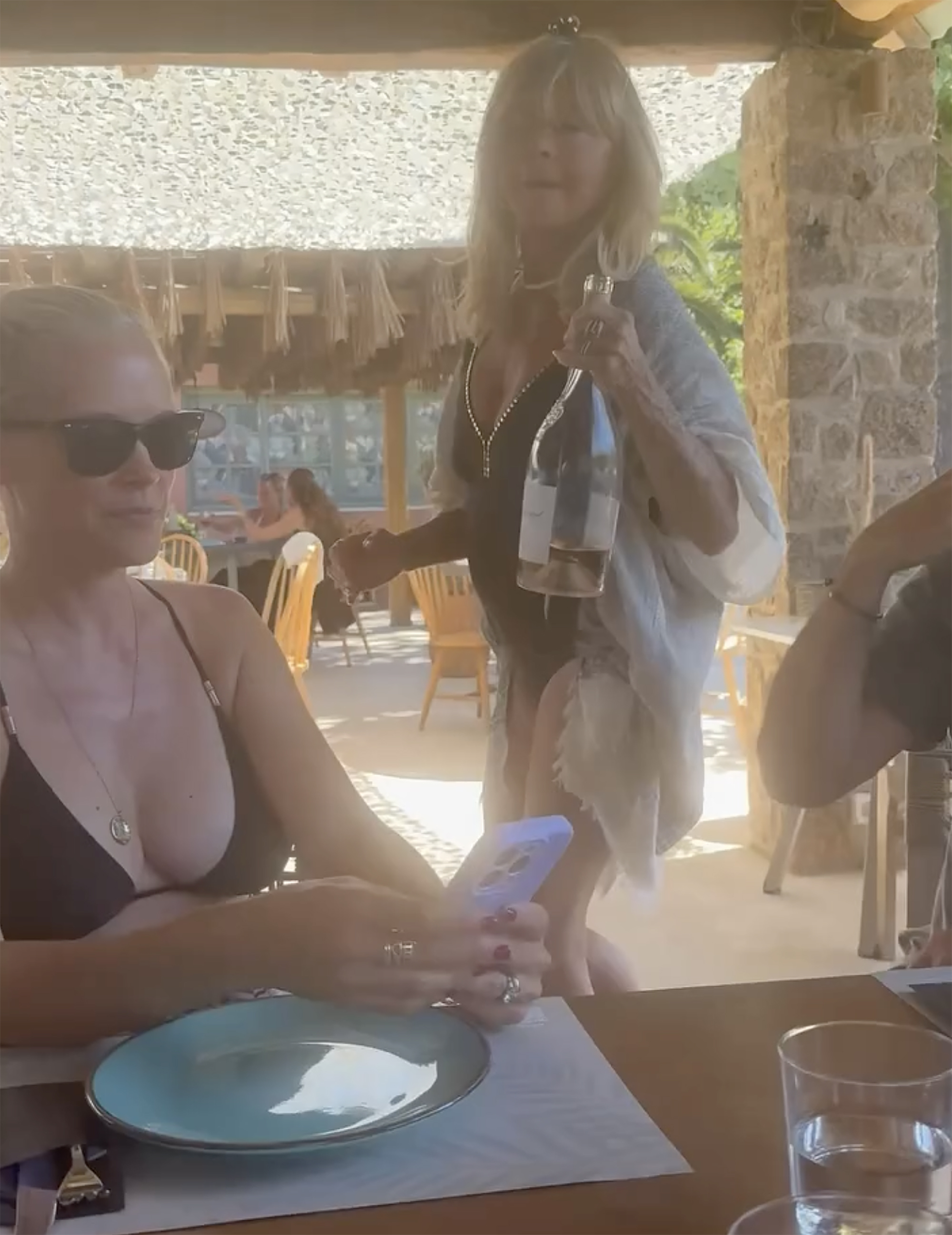 Goldie Hawn standing and holding a bottle of champagne while another woman sits at a table.