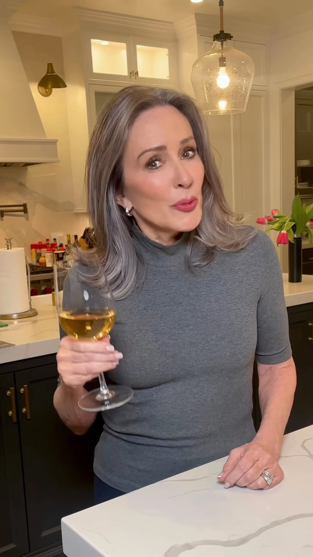 Patricia Heaton holding a glass of white wine while standing in a kitchen.