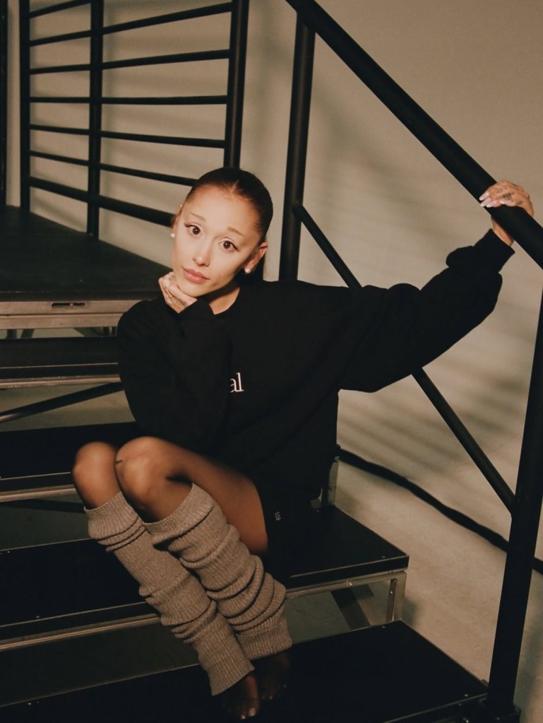 Ariana Grande in a black sweatshirt, black shorts, and gray leg warmers, sitting on stairs with her hand on her chin.