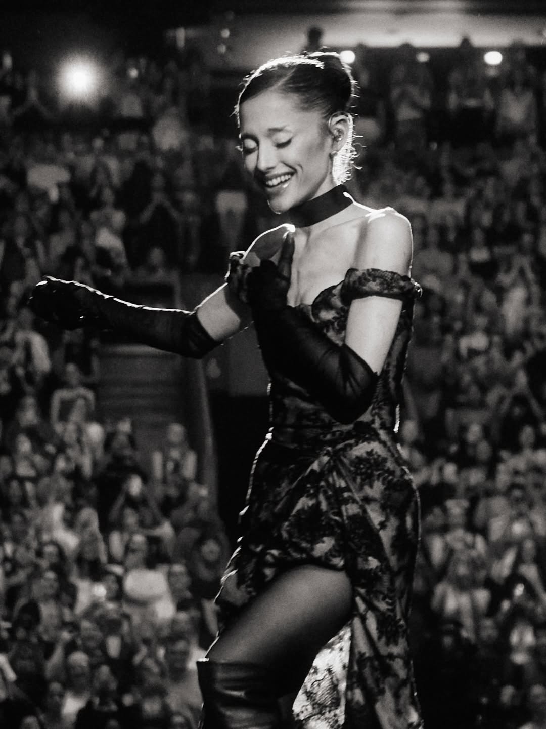 Ariana Grande smiling on stage with a crowd in the background.