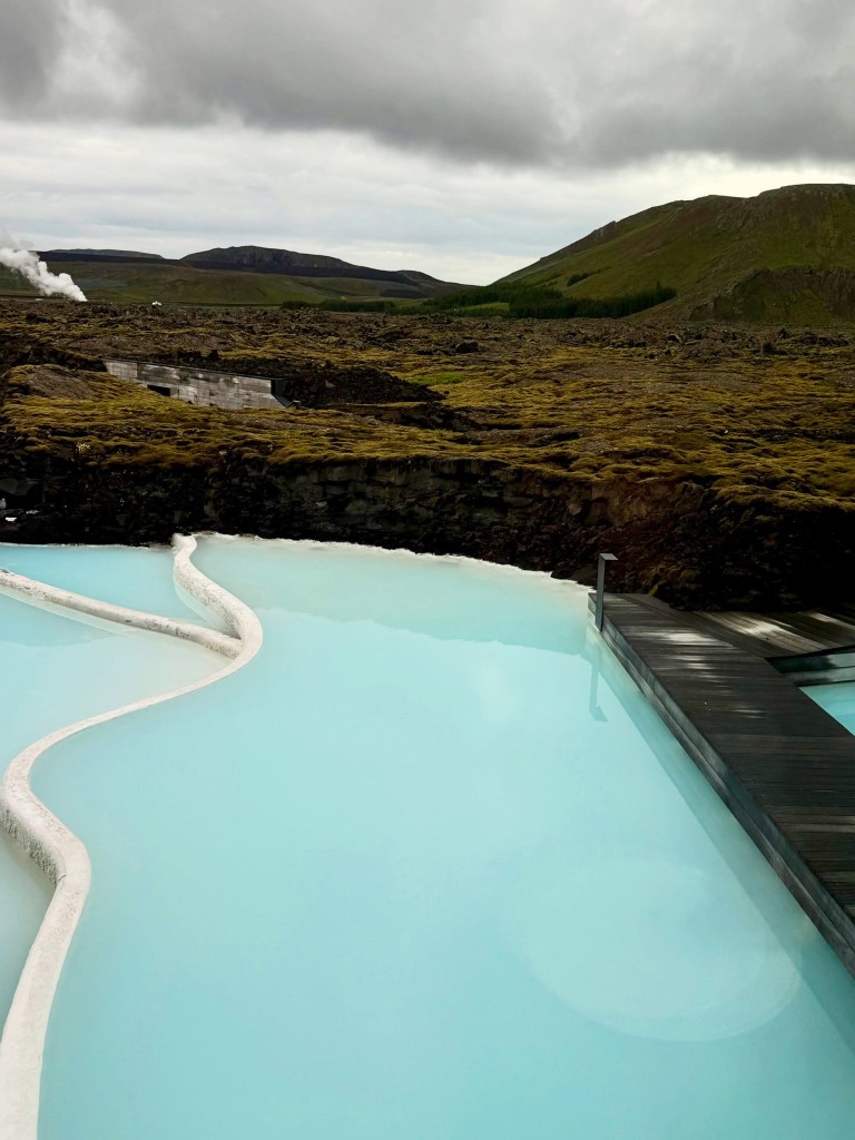 The Blue Lagoon geothermal spa in Iceland with a steamy industrial building and green hills in the distance.
