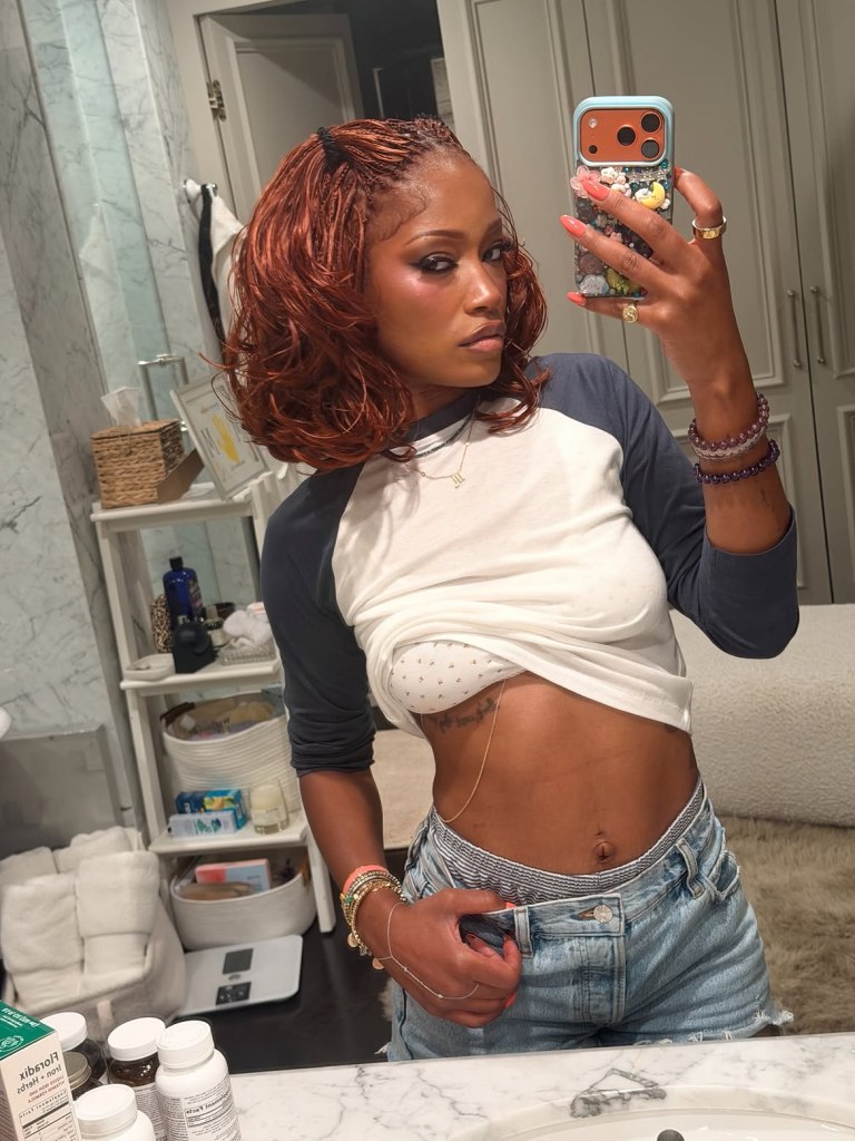 Keke Palmer poses for a mirror selfie in a bathroom, wearing a white and blue baseball tee, denim shorts, and a bra.