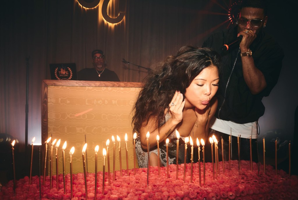 Jen Rubio blowing out candles on a raspberry cake with Nelly in the background.