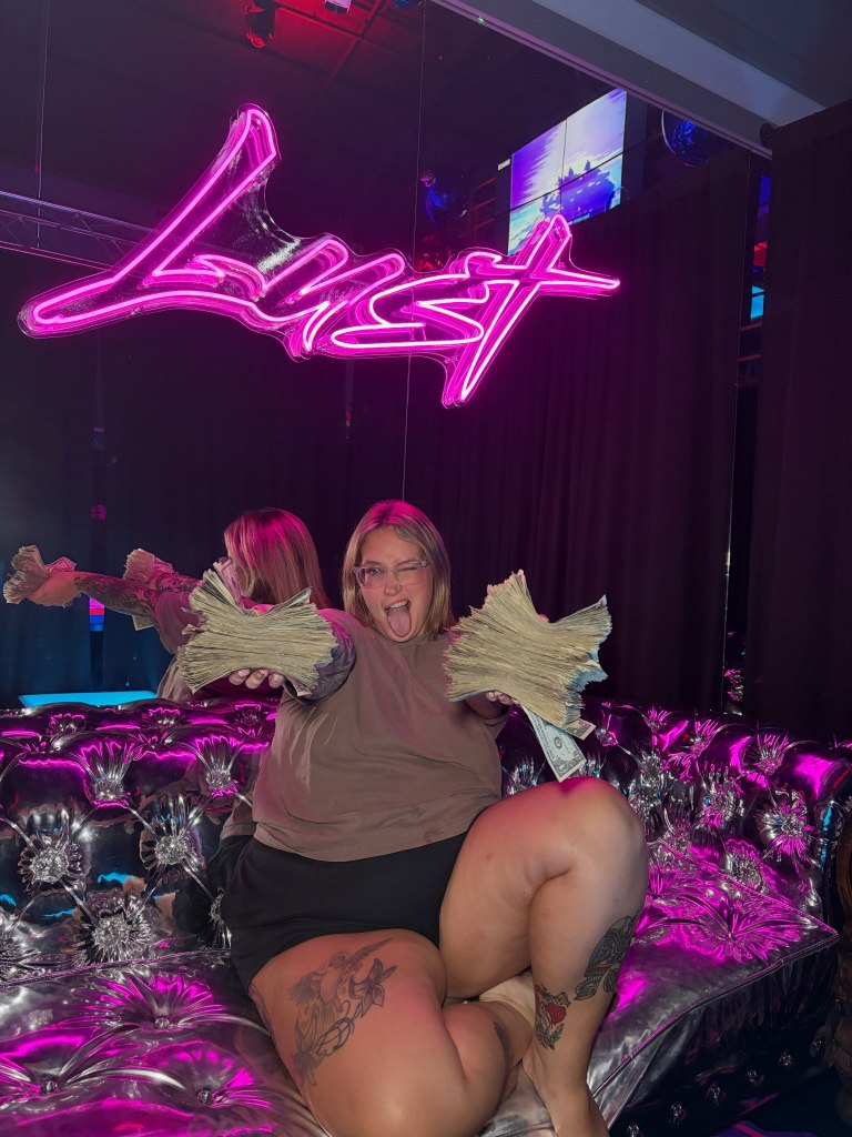 A woman holding fanned-out stacks of money on a couch with a neon sign above her head.