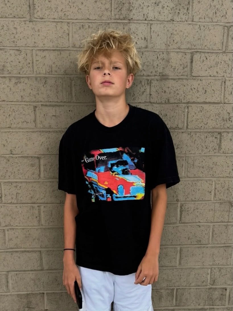 Axl Duhamel, son of Fergie and Josh Duhamel, wearing a black t-shirt with a colorful car graphic and white shorts, against a brick wall.