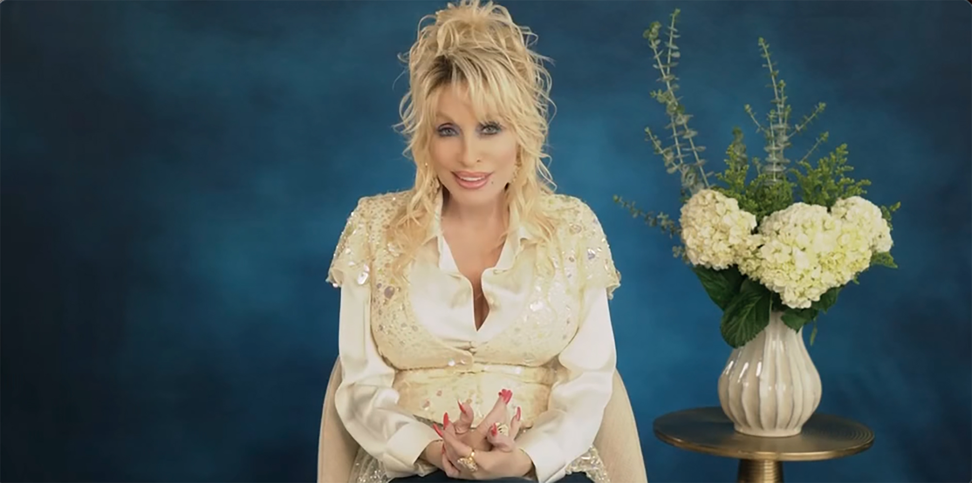 Dolly Parton seated against a blue background, wearing a sequined top.