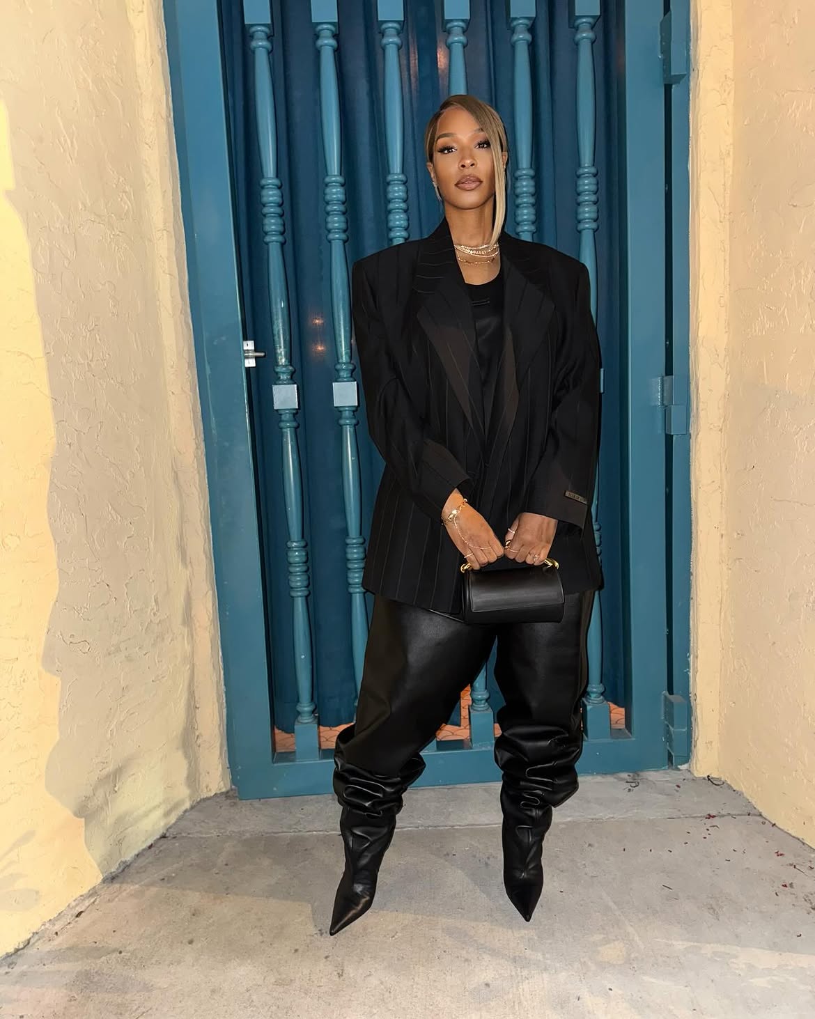 Savannah James in a black pinstripe blazer, black top, black leather pants, and black pointed-toe boots, holding a small black handbag.