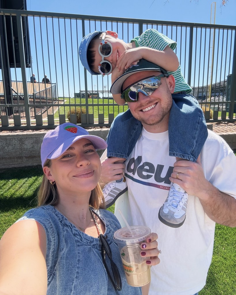 A family selfie shows Emilie Kiser holding a drink, her husband smiling with their child on his shoulders, all outside in front of a fence and field.