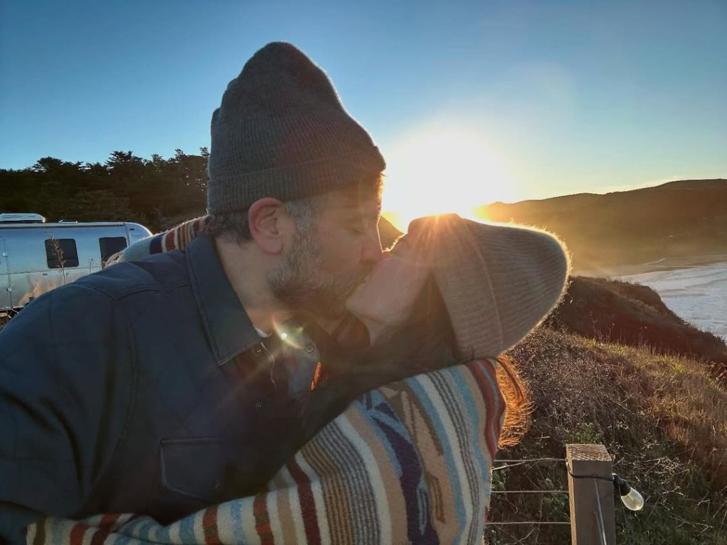 Josh Radnor and his wife kissing at sunset by the ocean.