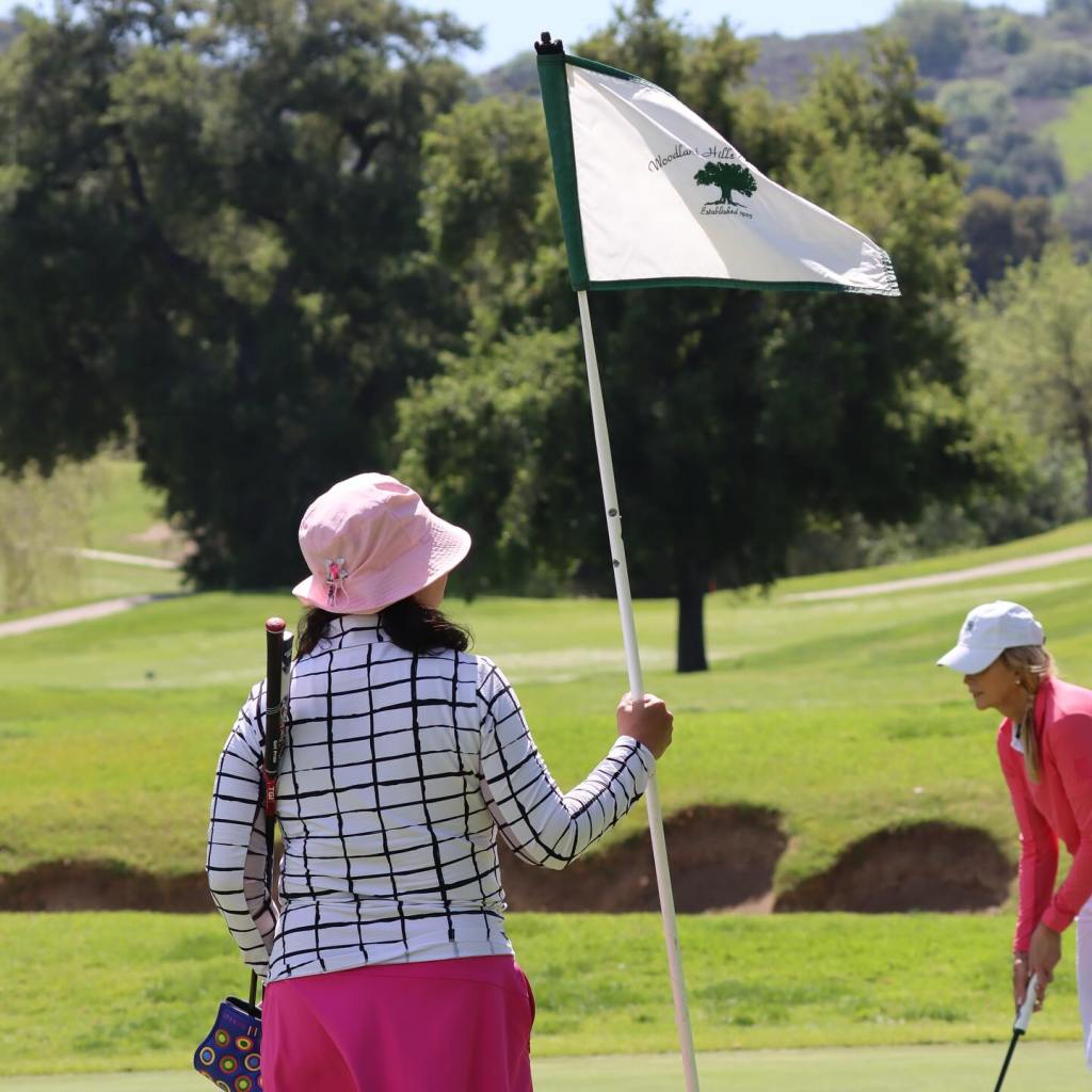 Two women on a golf course, one holding a flagstick from Woodland Hills, the other preparing to putt.