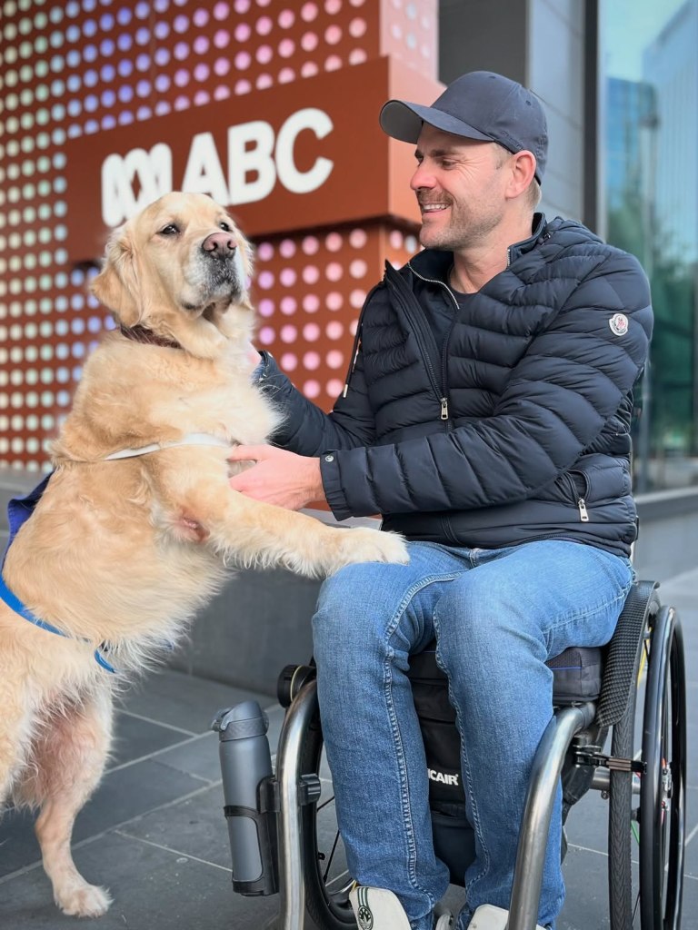 Shane Hryhorec in a wheelchair with his golden retriever service dog, with an ABC sign in the background.