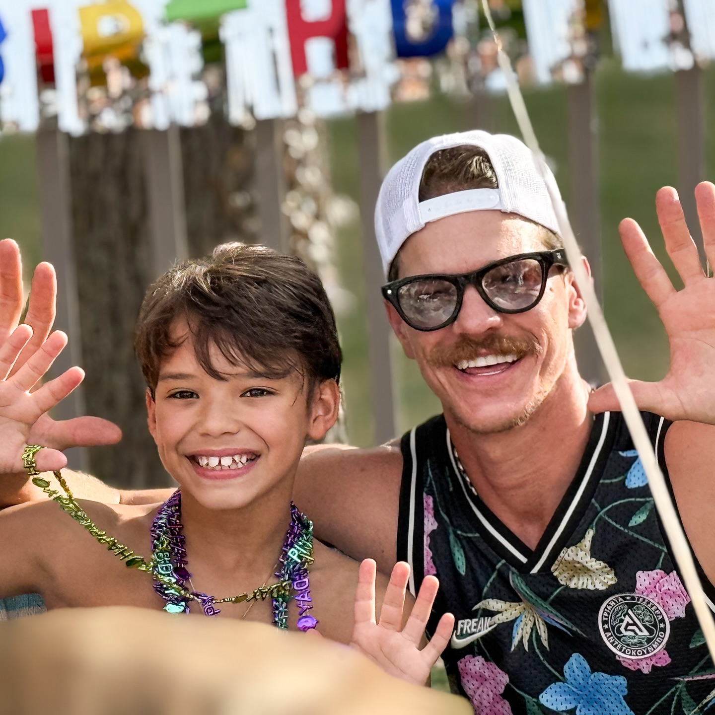 Josey and Ryan Dorsey in beaded necklaces with an adult man in glasses and a backwards cap, both waving their hands.