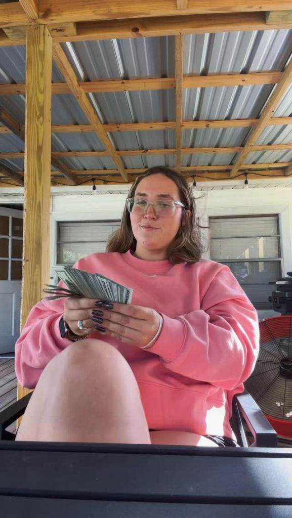 A young woman in a pink sweatshirt sitting on a porch counting a fan of money.
