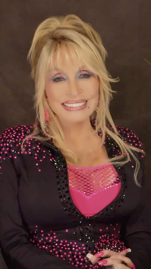 Dolly Parton smiling, wearing a black jacket with pink sequins over a hot pink top with a mesh neckline.