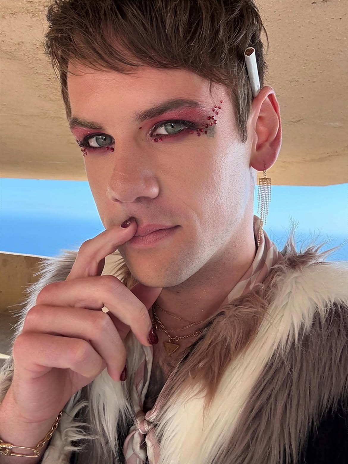 Caleb Shomo with red eyeshadow and glitter, a cigarette behind his ear, and a finger to his painted lips.