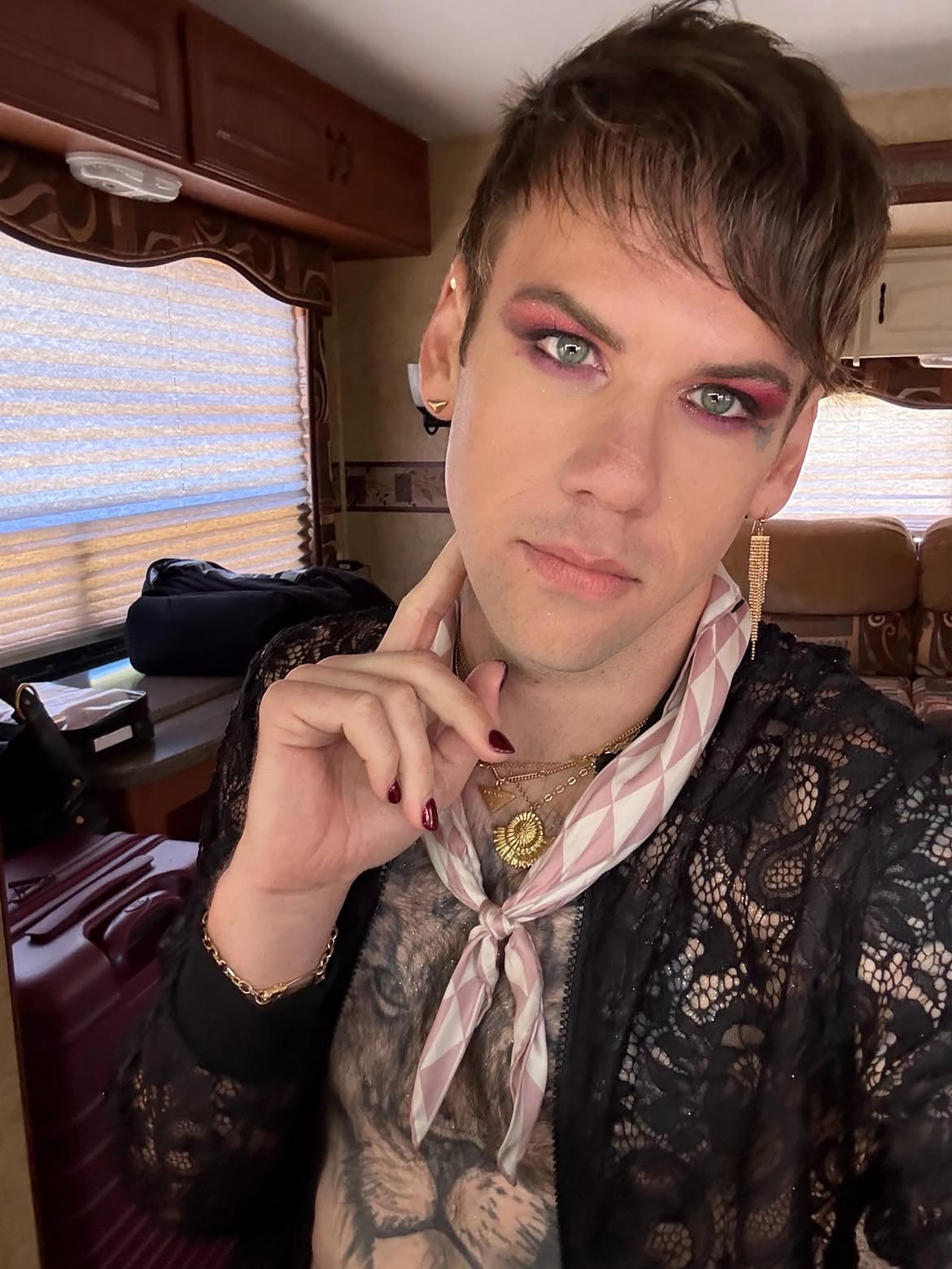 Caleb Shomo wearing full makeup, earrings, necklace, and a patterned scarf, looking at the camera.