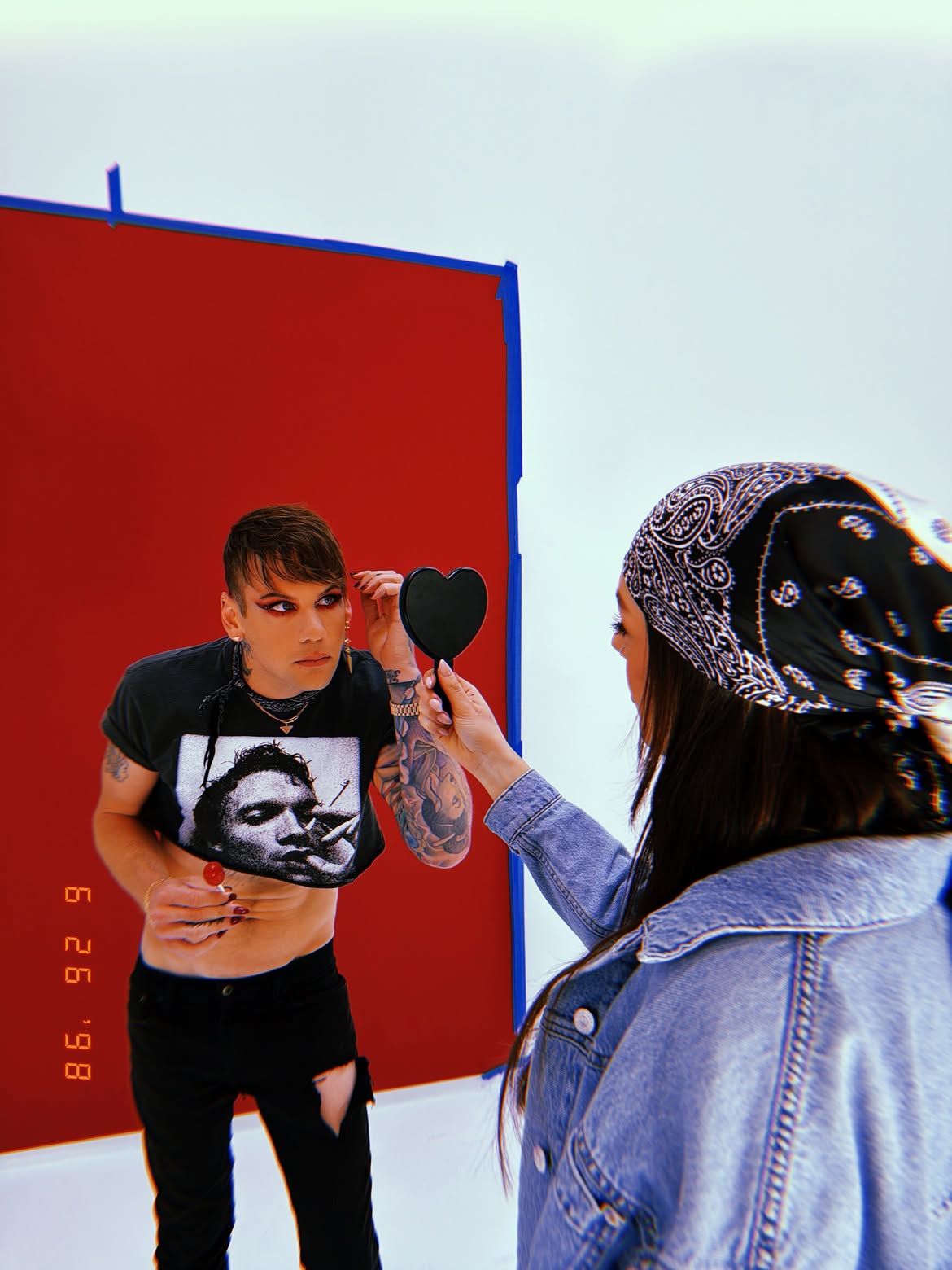 Caleb Shomo applying makeup while looking into a heart-shaped mirror held by another person.