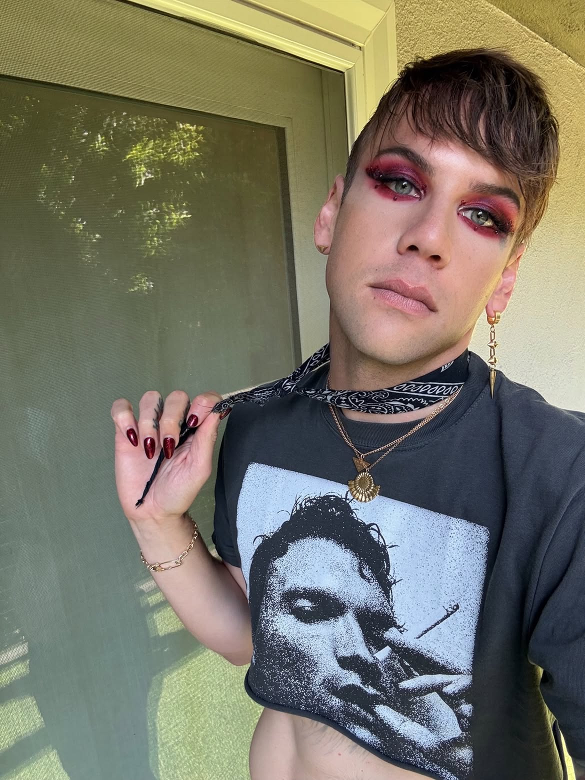 Caleb Shomo with red eye makeup and dark red nails holding a bandana, wearing a black crop top.