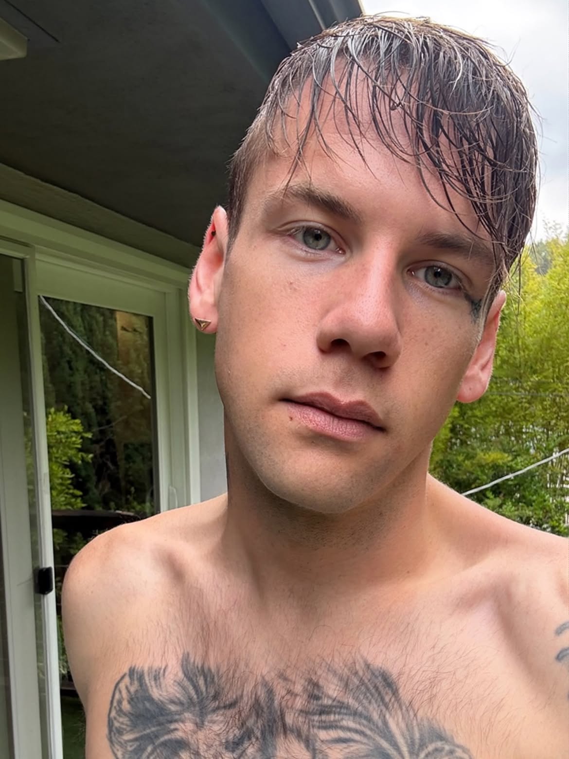 Caleb Shomo with wet hair and a chest tattoo, standing shirtless outdoors.