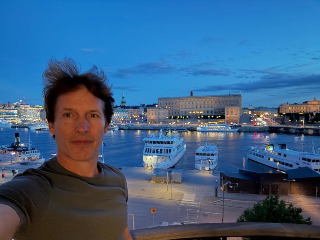 James Blunt in front of a European cityscape with boats docked along a river at dusk.
