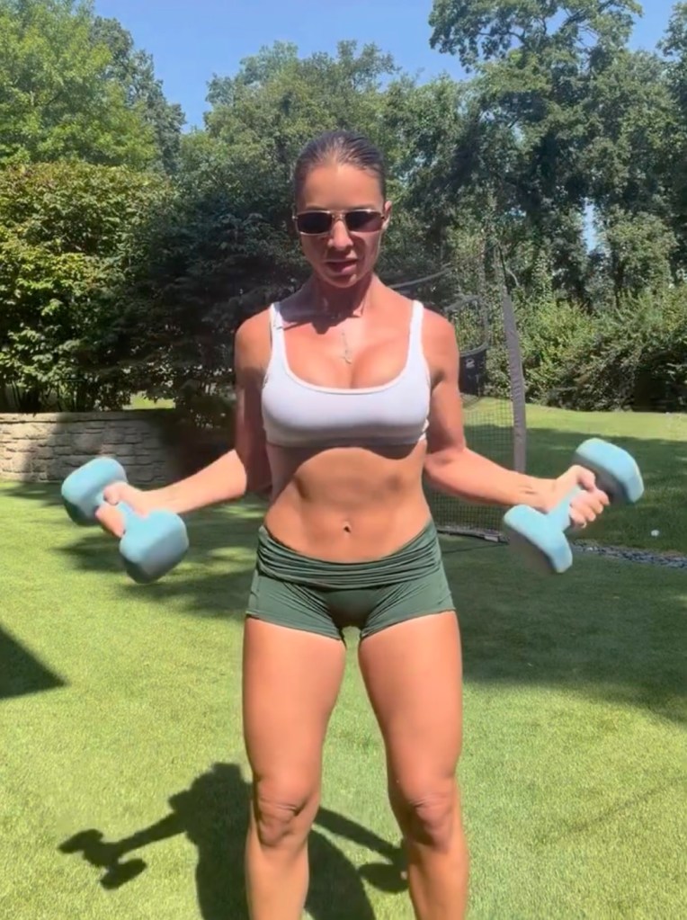 A woman in sunglasses and athletic wear exercising outdoors with dumbbells.