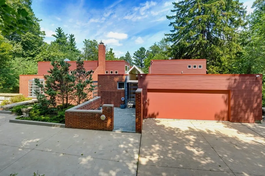 Modern red-brick house with a red garage door, nestled amongst green trees.
