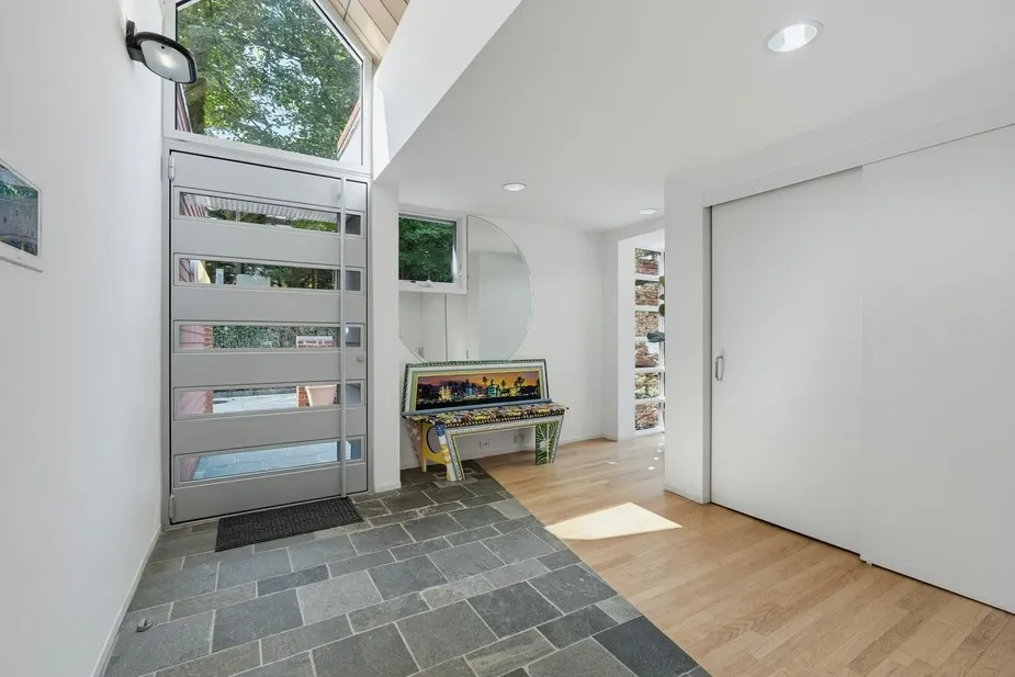 The entrance of an upside-down house by architect Kenneth Neumann, featuring slate tile flooring, a modern door with horizontal glass panels, and a colorful decorative bench.
