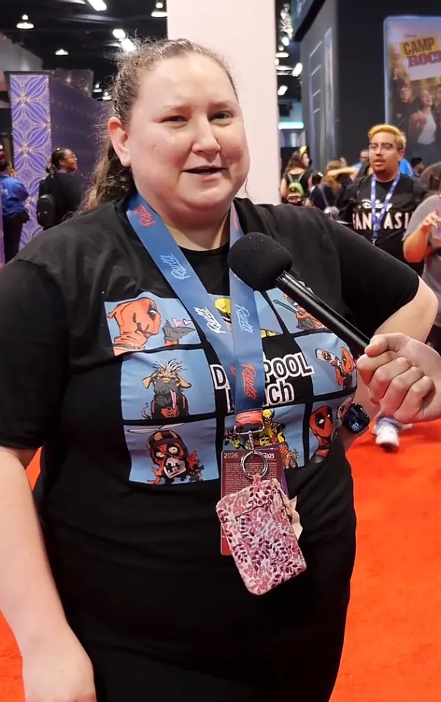 A woman in a black Deadpool shirt speaks into a microphone at D23.