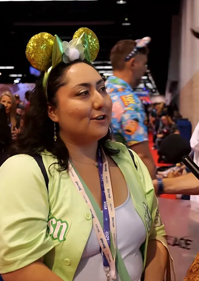 A young woman with green sequined Minnie Mouse ears on her head and a light green button-up shirt over a white tank top, talking to an interviewer.