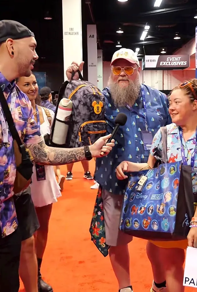Two men and two women at a Disney event with one man holding a Mickey Mouse backpack and another man holding a microphone towards him, while a woman holds a Disney-themed bag.