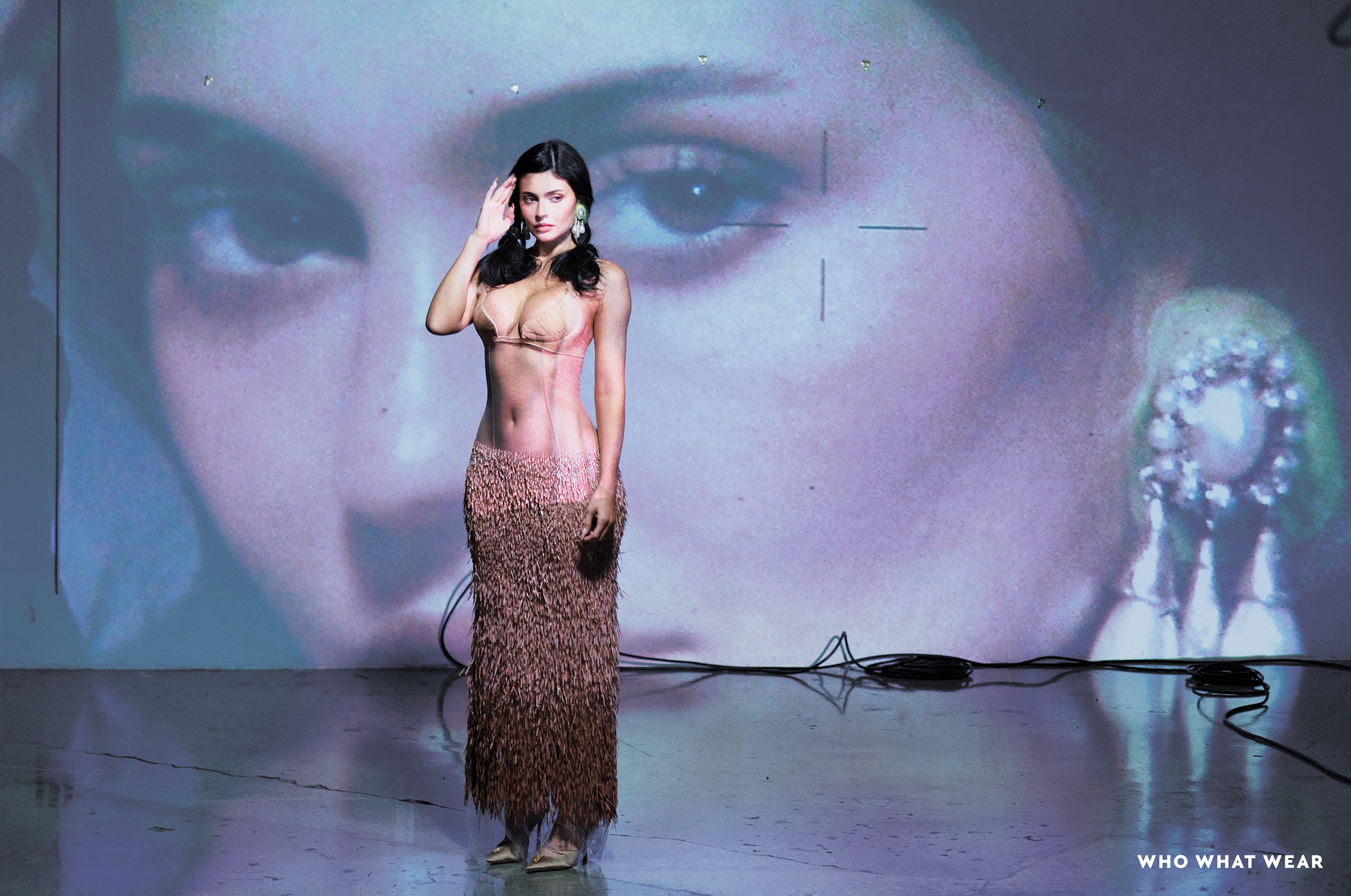 Kylie Jenner wearing a light pink bra and a shaggy peach-colored skirt, standing in front of a projection of a woman's eye.