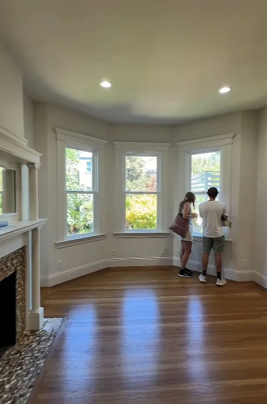 A man and a woman looking out bay windows in an empty room with hardwood floors and a fireplace.