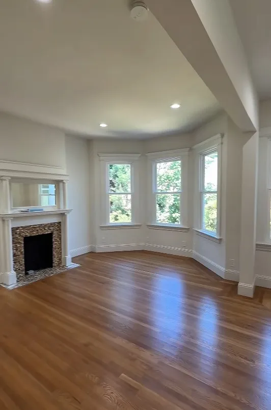 A view of an empty room with wood floors, bay windows, and a fireplace with a mirror above it.