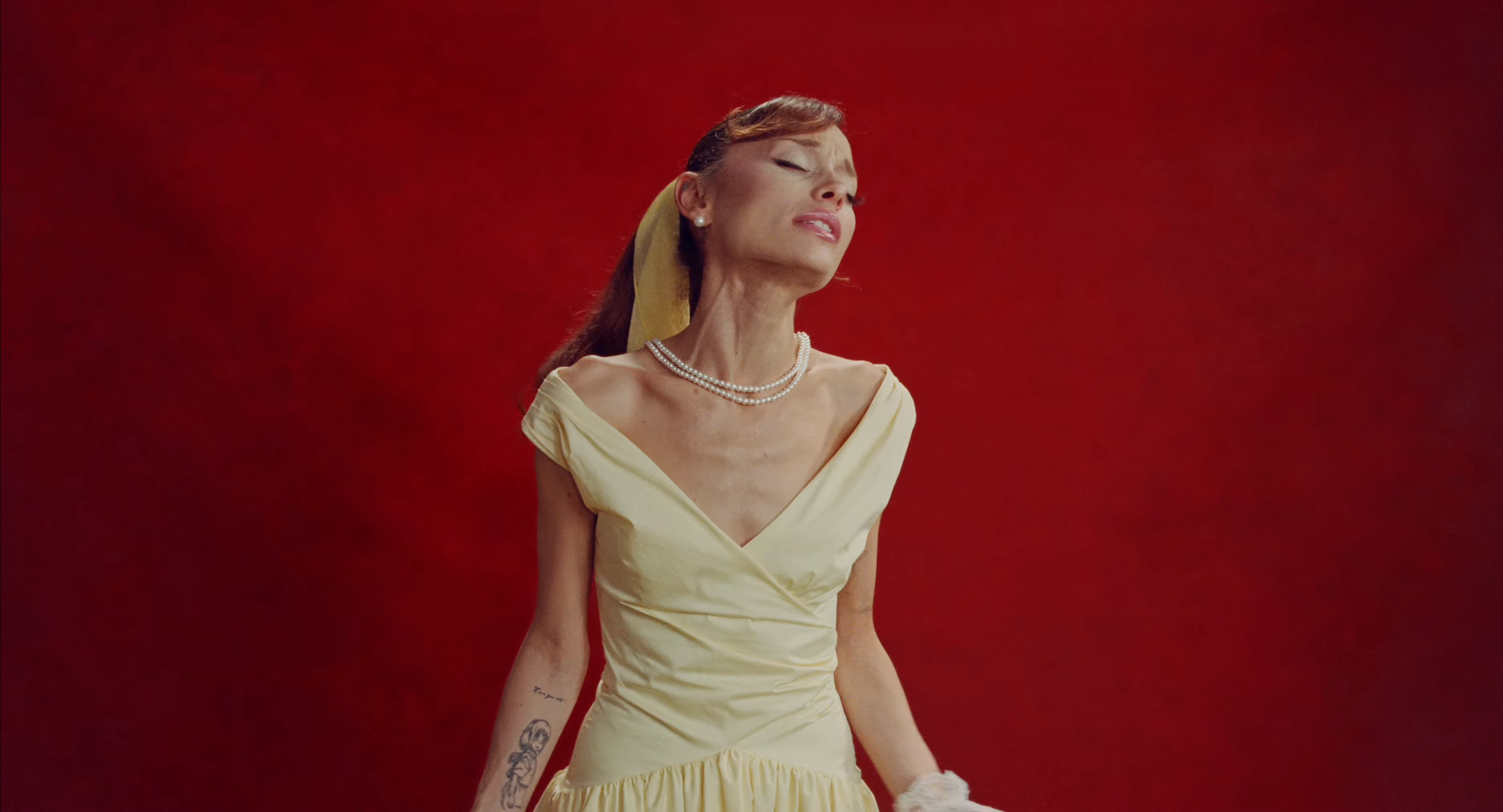 Ariana Grande in a yellow dress and pearl necklace, with a tattoo on her arm, against a red background.