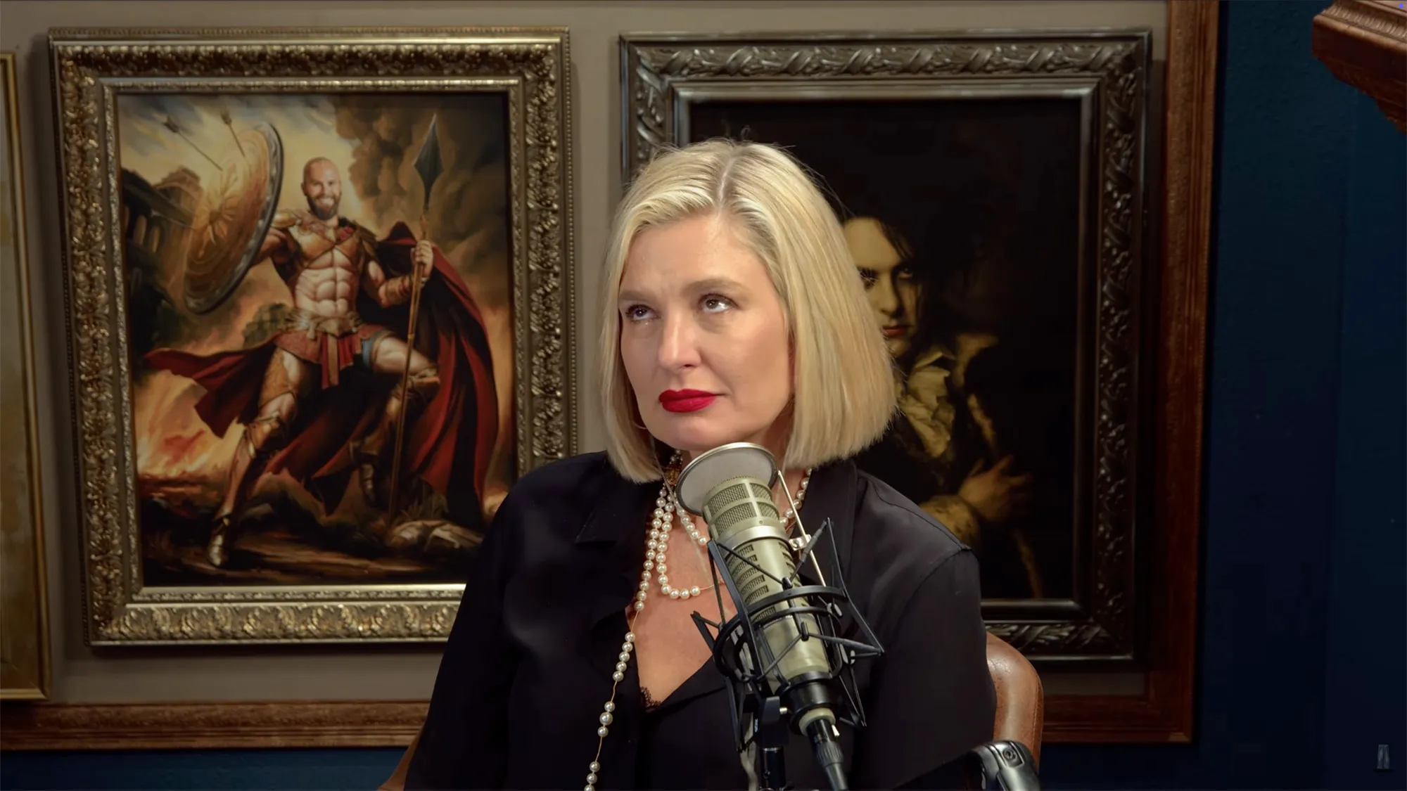 Christina Pazsitzky sits at a microphone with two paintings behind her.