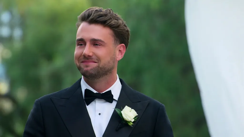 A man in a tuxedo with a white rose boutonnière and bow tie, with tears in his eyes, smiling.