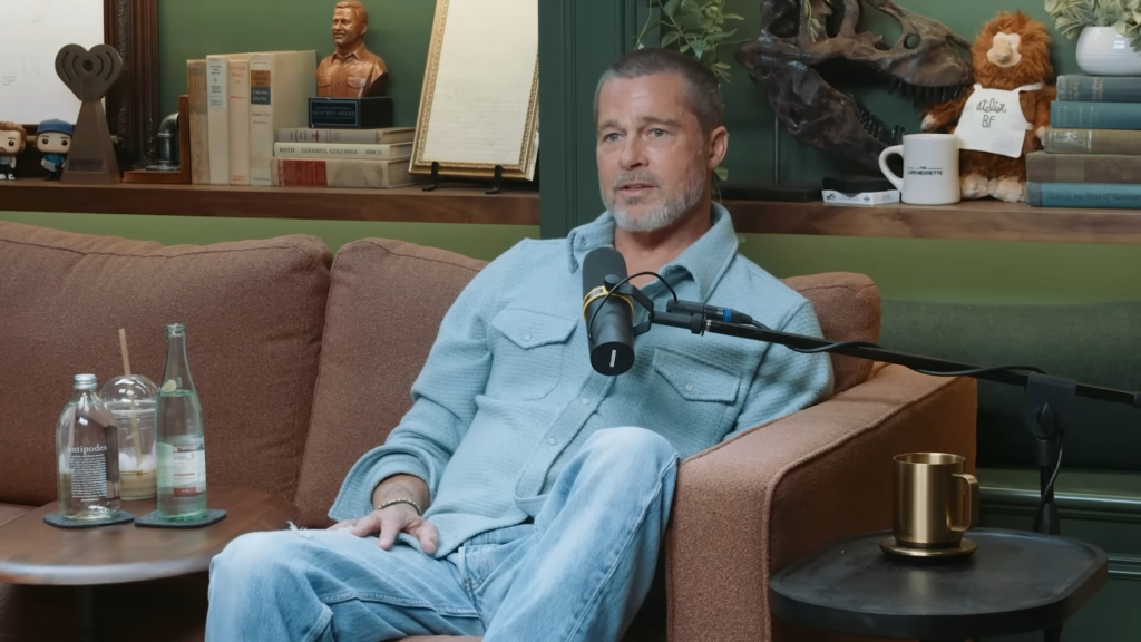 Brad Pitt sits on a brown sofa, wearing a light blue shirt and jeans, next to a microphone during an interview.