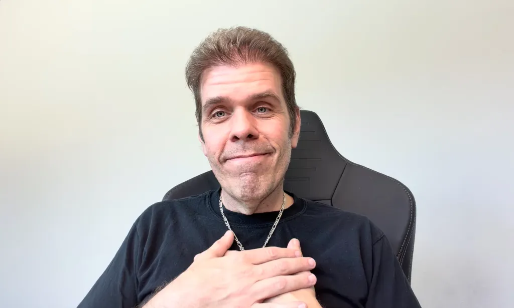 A man in a black shirt sitting in a black chair with his hands over his heart.