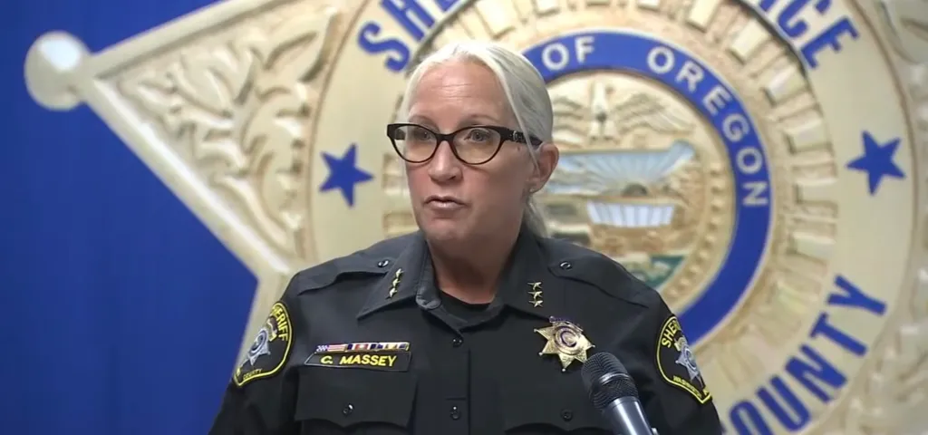 A sheriff in uniform, identified by her name tag as C. Massey, speaks at a podium with a large sheriff's star emblem behind her.